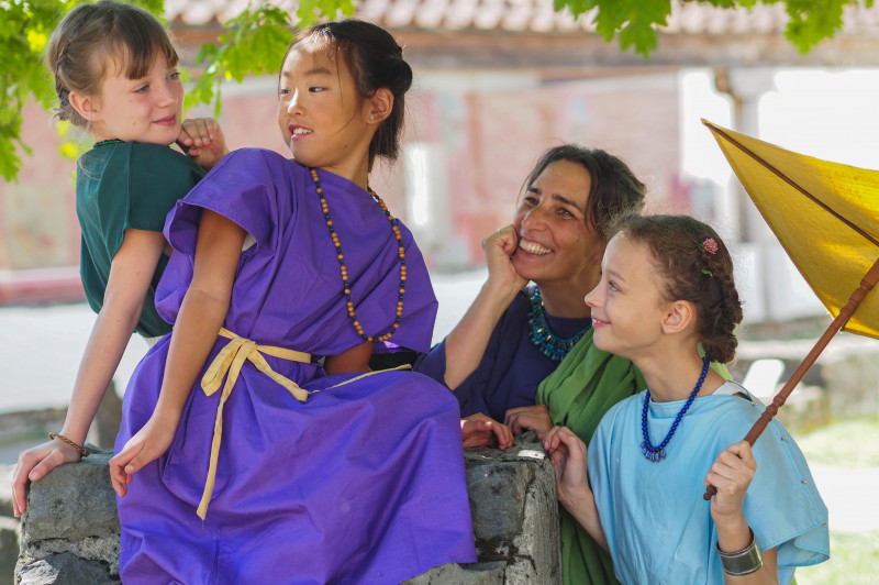 Drei Mädchen und eine Frau in römischer Kleidung stehen im Tempelbezirk des APC vertraut beieinander