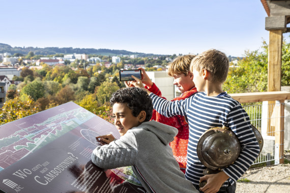 Drei Jungen stehen an Infotafel und blicken von Aussichtspunkt im APC auf die Stadt Kempten