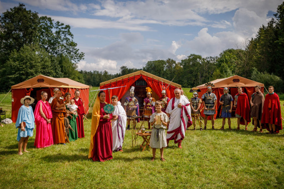 Darsteller:innengruppe in historischen, römischen Kostümen auf einer Wiese vor 3 Zelten