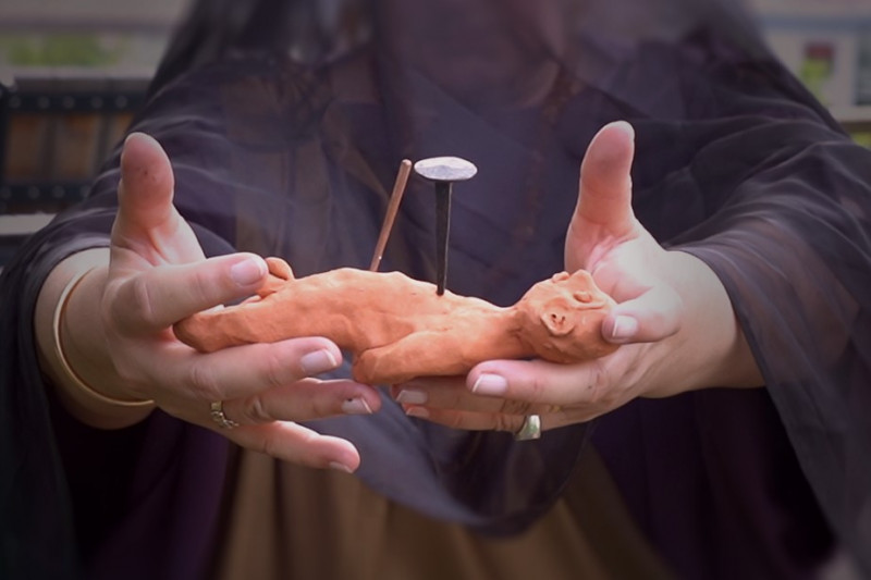 Eine verschleierte Frau hält eine Tonpuppe eines Menschen in den Händen, in der ein Nagel steckt