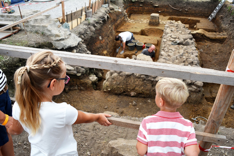 Zwei Kinder blicken in eine Ausgrabung, es sind Archäologen bei der Arbeit sichtbar